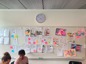 a whiteboard coverd in post-it notes and white pieces of paper. a clock above, two people concentrating in the bottom left corner of the shot.