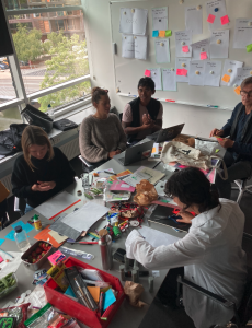 a long table covered in pens, paper , boxes, zines, glue, paper, scizzors. five pepole sit around it, two on laptops, the others working on A4 pages. there is an atmosphere of activity