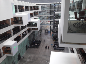 looking into the atrium of the IT University of Copenhagen, one sees 'boxes' (rooms) jutting out of the side, and people wandering around far below. The photo is taken near the ceiling, where light is streaming in.